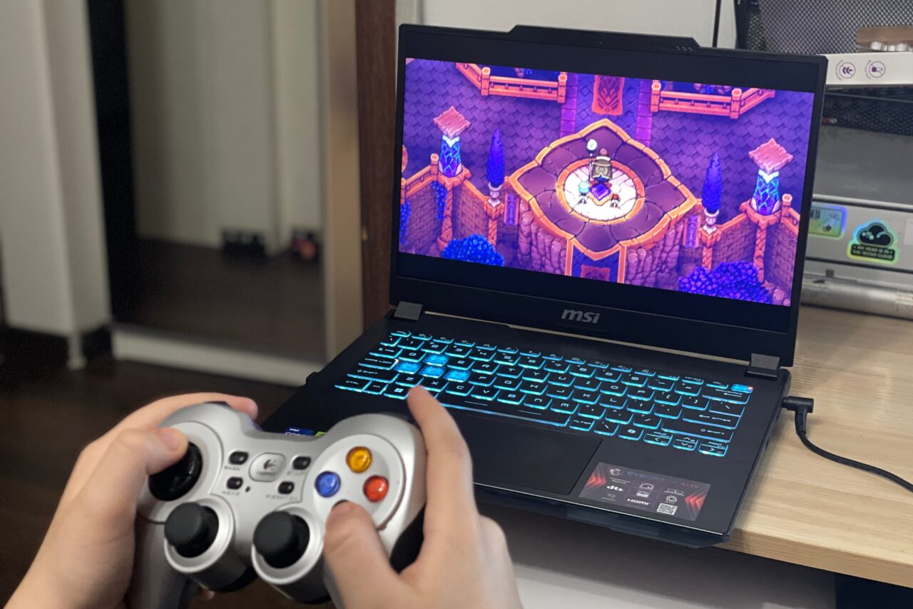 Dłonie trzymające bezprzewodowy kontroler do gier przed laptopem marki MSI z włączoną grą o estetyce piksel art na ekranie. Recenzja MSI Cyborg 14. 