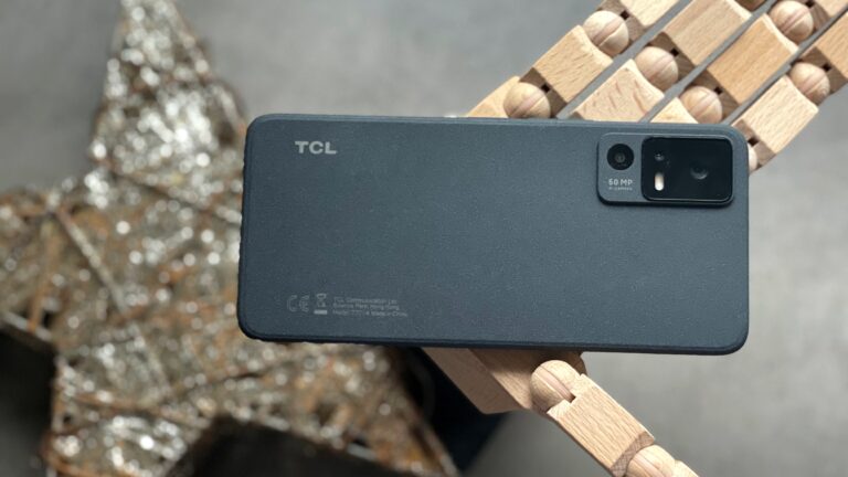 Telefon komórkowy marki TCL z aparatem 50 MP umieszczony na drewnianej powierzchni, na tle rozmytej metalicznej dekoracji.