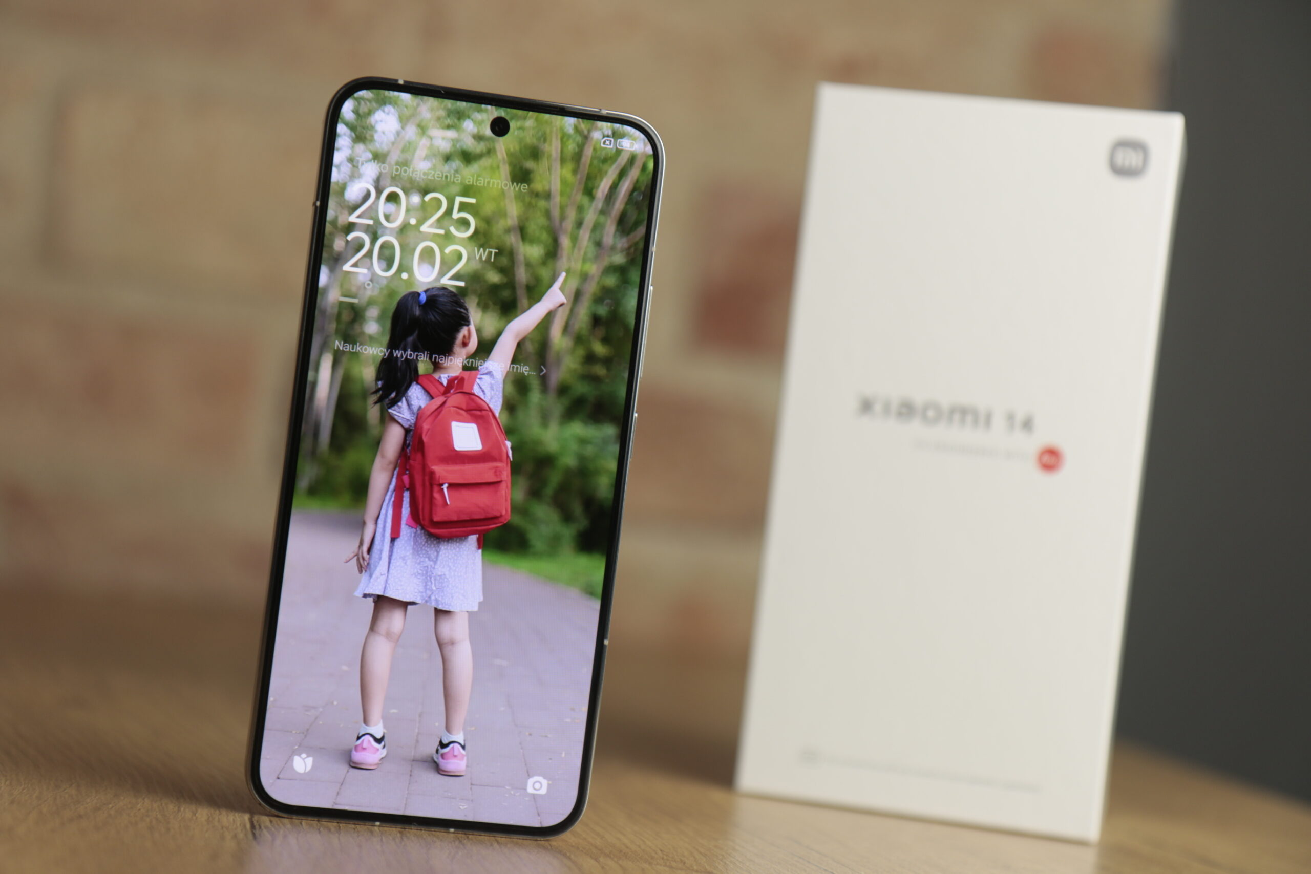 Recenzowany smartfon Xiaomi 14 z wyświetlanym na ekranie obrazem małej dziewczynki z plecakiem oraz opakowanie testowanego produktu w tle.
