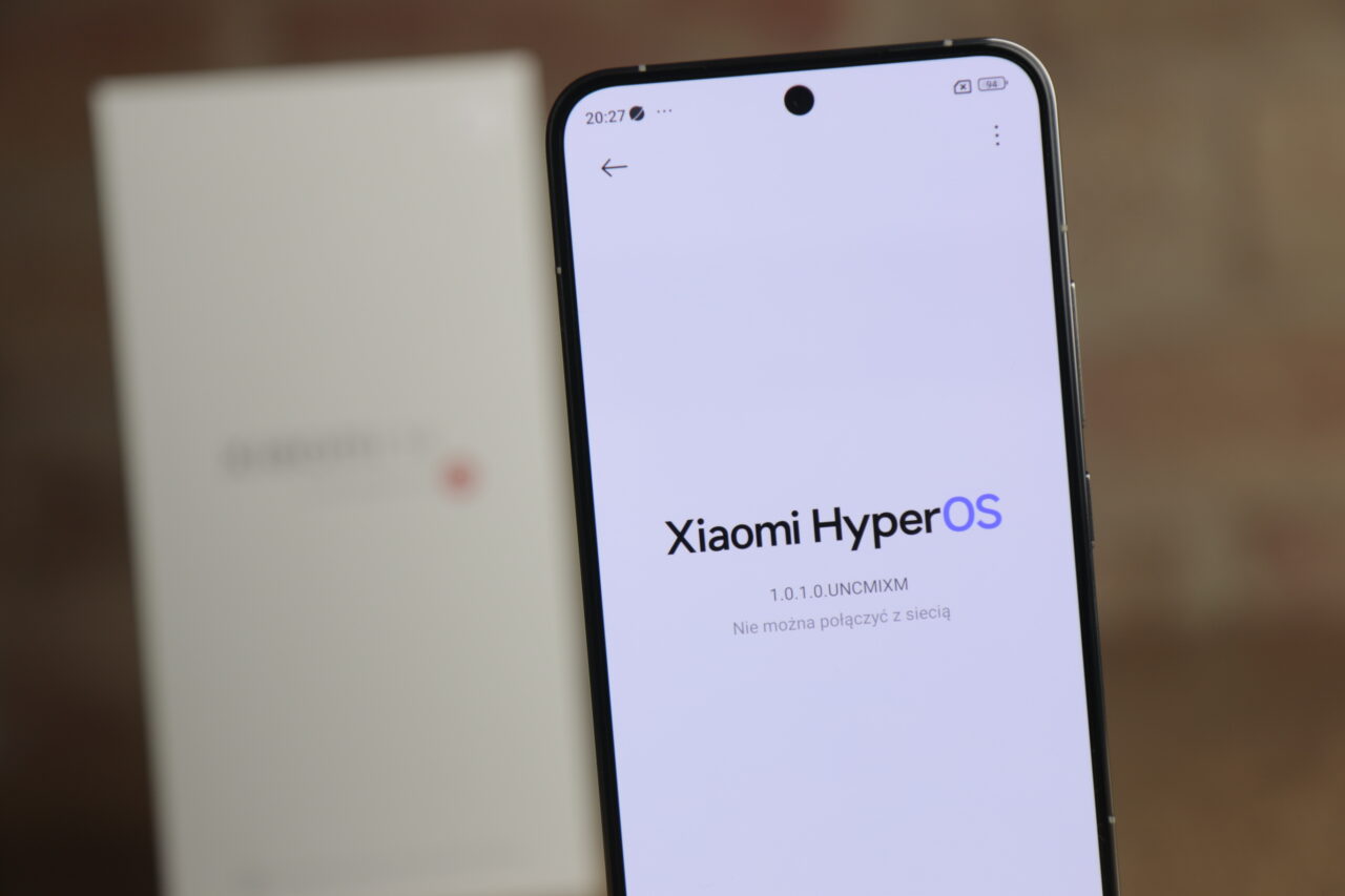 Testowany smartfon Xiaomi 14 wyświetlający ekran startowy z napisem "Xiaomi HyperOS" na tle rozmytej białej pudełkowej obudowy.