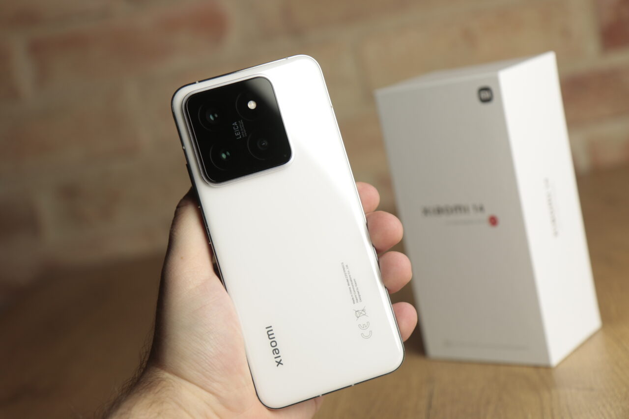 Tylna strona białego smartfona z aparatem Leica trzymanego w dłoni, z opakowaniem w tle.
