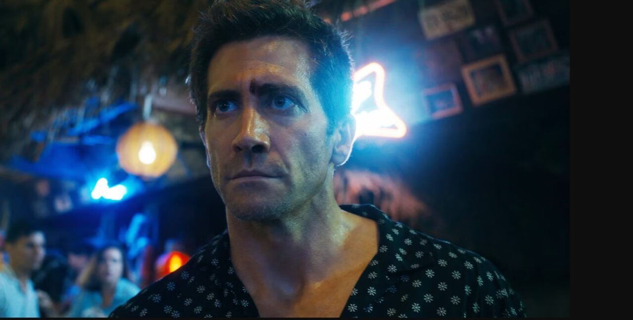 Kadr z filmu Road House. Mężczyzna o poważnym wyrazie twarzy w ciemnym pomieszczeniu z neonowymi światłami w tle.