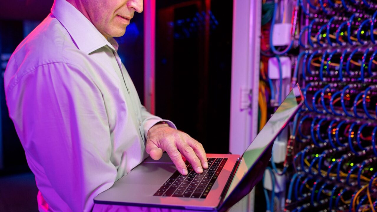 Mężczyzna w białej koszuli pracujący na laptopie z czerwoną klapą, w tle serwerownia z aktywnym sprzętem sieciowym.