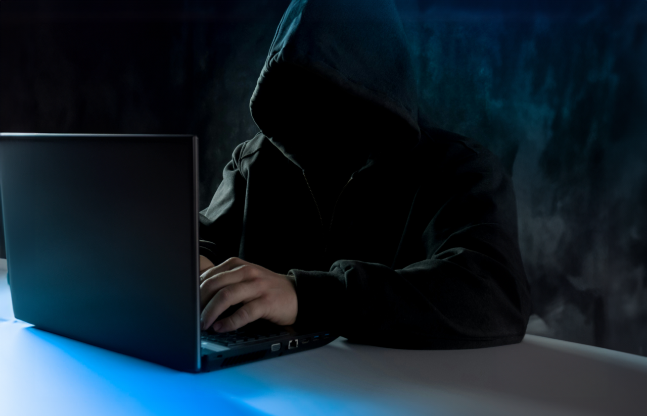 Osoba w kapturze pracująca na laptopie w słabo oświetlonym pomieszczeniu, emitującym niebieskie światło.
