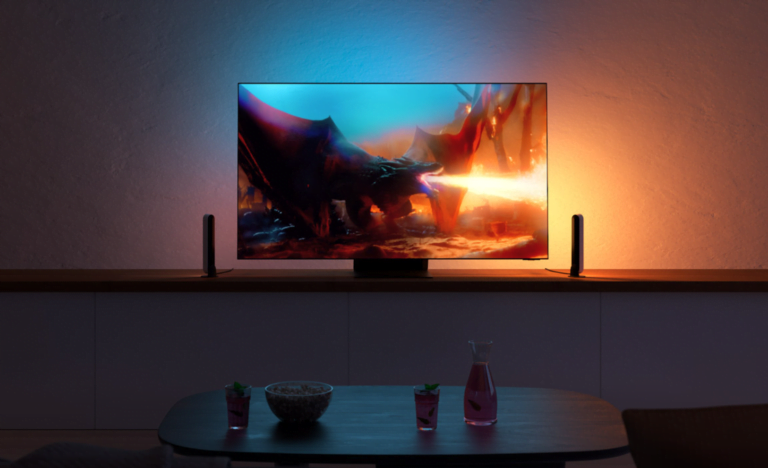 Telewizor na komodzie wyświetlający scenę z filmu z smokiem wydychającym ogień, przed nim stół z misą popcornu oraz dwoma kolorowymi napojami.