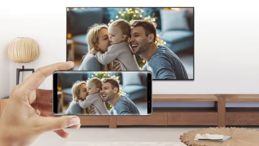 Mão segurando um smartphone que transmite a imagem de uma família feliz em uma TV ao fundo, em um ambiente tranquilo.