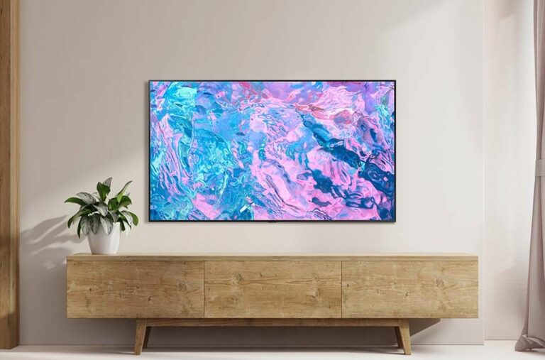 Telewizor 4K zawieszony na ścianie wyświetlający abstrakcyjny, kolorowy obraz, umieszczony nad drewnianą komodą z rośliną w białym doniczce po lewej stronie i zasłoną po prawej w jasnym pokoju.