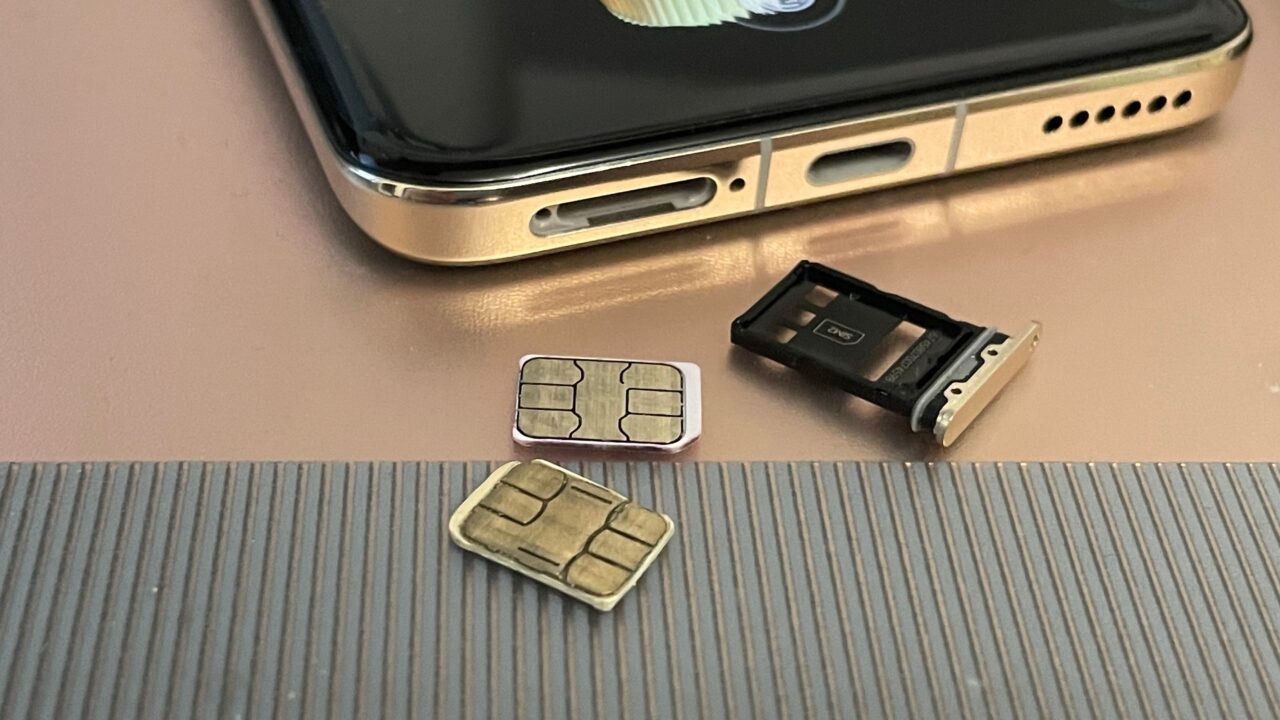 Klonowanie kart SIM. SIM swap. Złoty smartfon z wyciąganą tacką na kartę SIM, obok której znajdują się dwie karty SIM – jedna nowa i jedna zużyta – na szarej powierzchni z wąskimi rowkami.