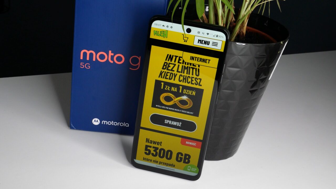 Smartfon marki Motorola wyświetlający reklamę oferty internetowej, obok pudełko z napisem Moto 5G i doniczka z rośliną.