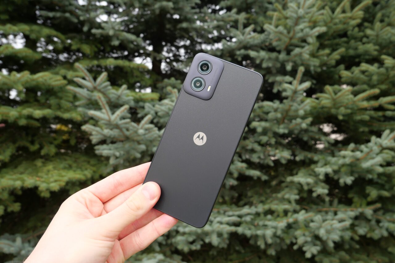 Ręka trzymająca smartfon Motorola z podwójnym aparatem fotograficznym na tle zielonych gałęzi drzew iglastych.