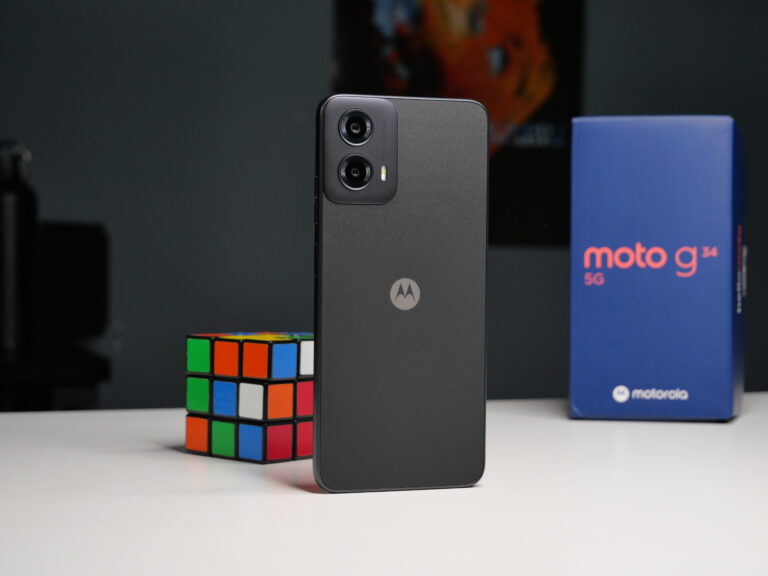 Czarny smartfon Motorola z podwójnym aparatem umieszczony pionowo na białym blacie, obok kostki Rubika i niebieskiego pudełka z napisem "moto g 5G".