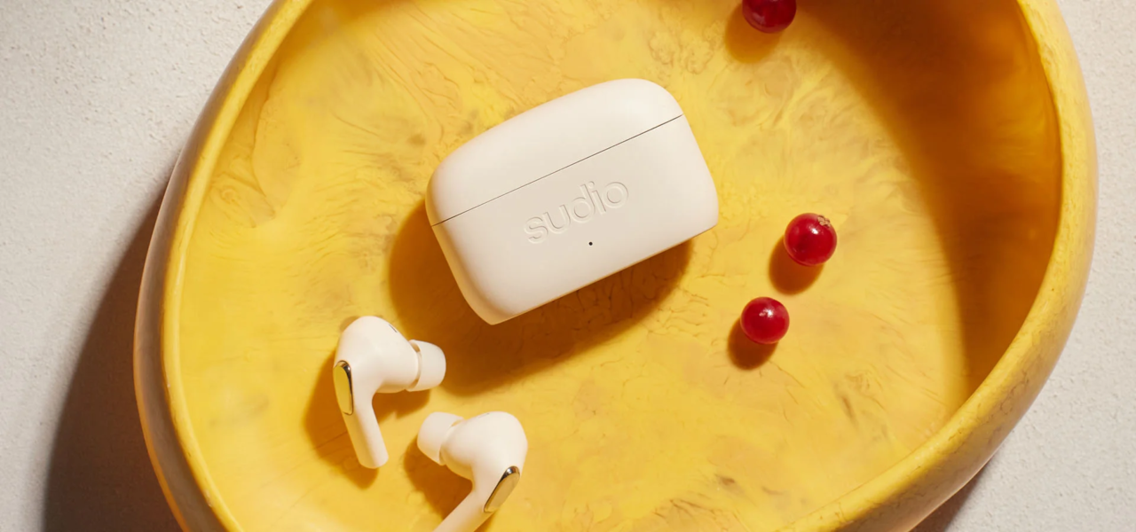 Białe bezprzewodowe słuchawki Sudio E3 i etui z napisem "sudio" na żółtym talerzu obok trzech czerwonych jagód.