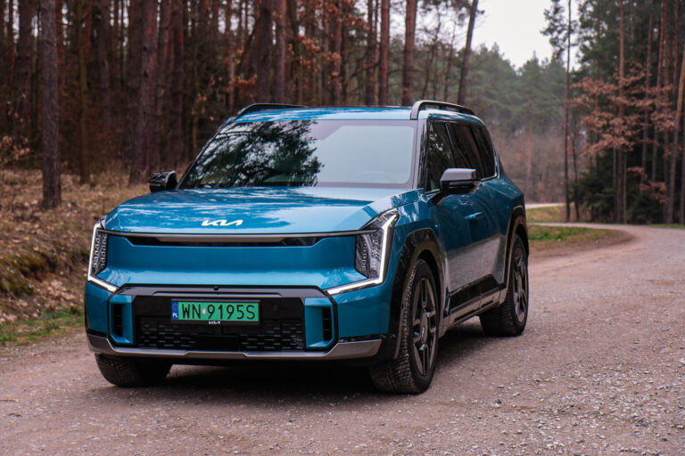 Niebieski SUV marki Kia EV9 zaparkowany na leśnej drodze żwirowej w ramach testu i wydania opinii.