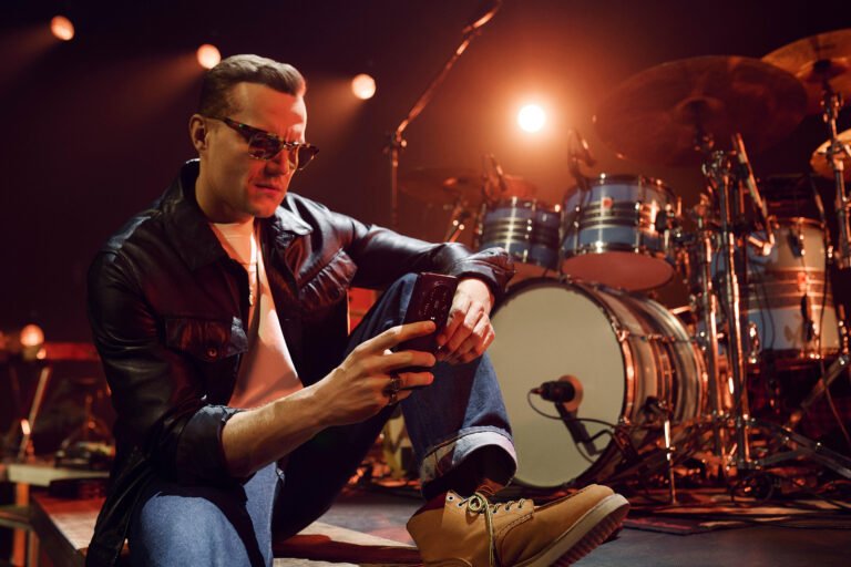 Mężczyzna w czarnej kurtce skórzanej i okularach przeciwsłonecznych siedzi na scenie z nogą założoną na kolano, przeglądając telefon, z perkusją w tle.