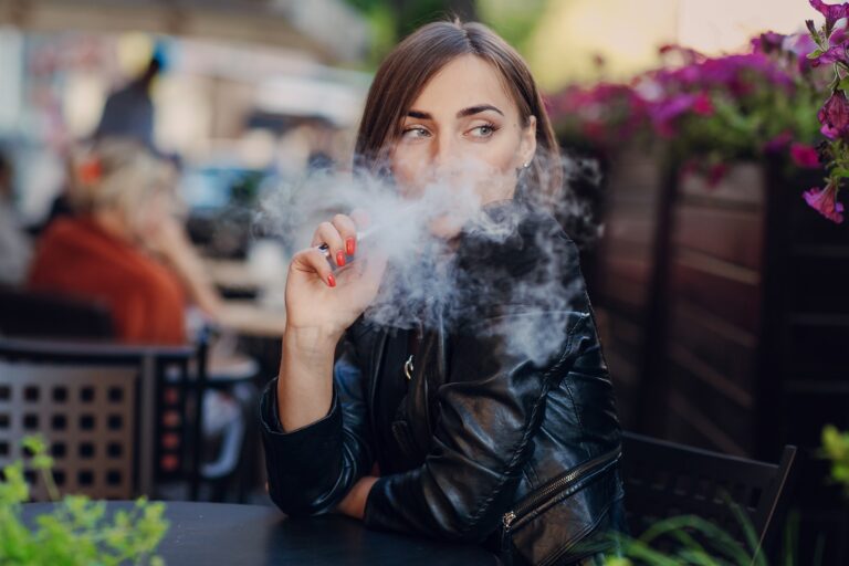 Kobieta w czarnej skórzanej kurtce pali papierosa na zewnątrz przy stoliku z kwiatami w tle.