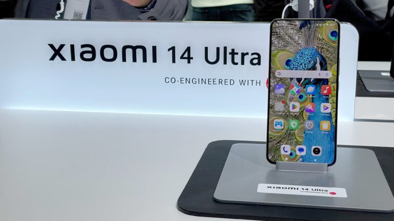 Smartfon Xiaomi 14 Ultra na wystawie, z ekranem wyświetlającym tapetę z pawiem, umieszczony na podstawce z logo producenta.