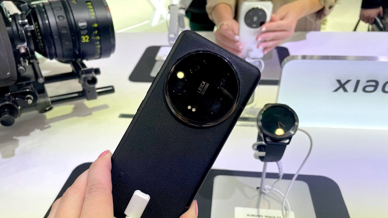 Osoba trzyma smartfon z obiektywem kamery Leica, w tle inne urządzenia elektroniczne prezentowane na stoisku z napisem "Xiaomi".
