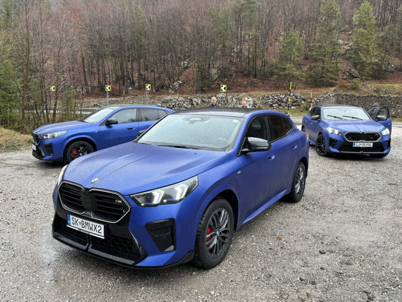 Trzy niebieskie samochody SUV zaparkowane jeden za drugim na żwirowym parkingu z drzewami w tle i dwoma osobami stojącymi przy najdalszym pojeździe.