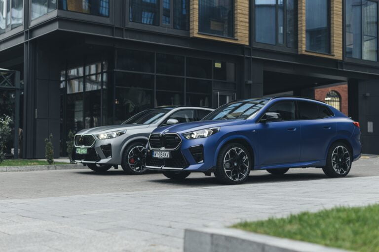 Dwa samochody BMW SUV zaparkowane obok siebie, przed nowoczesnym budynkiem z dużymi oknami, jeden koloru niebieskiego a drugi szarego.