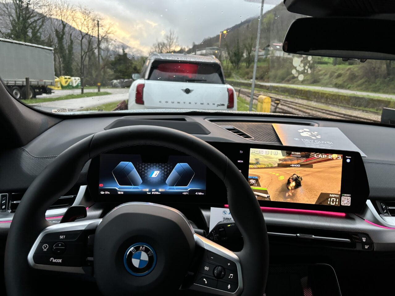 Widok z wnętrza samochodu marki BMW w kierunku deszczowej szyby przedniej, z kierownicą i wyświetlaczem deski rozdzielczej na pierwszym planie, na którym wyświetlana jest gra wyścigowa, w tle inne pojazdy i zielony krajobraz.