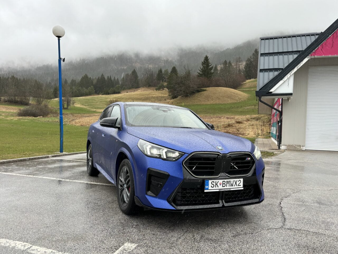 Niebieski samochód marki BMW zaparkowany na mokrym parkingu z widokiem na zielone wzgórza i gęstą mgłę w tle.