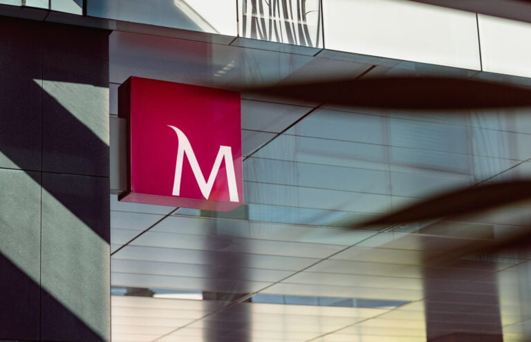 Różowy znak z logo Banku Millennium na metalowym wsporniku, umieszczony na tle nowoczesnej, szklanej fasady budynku.