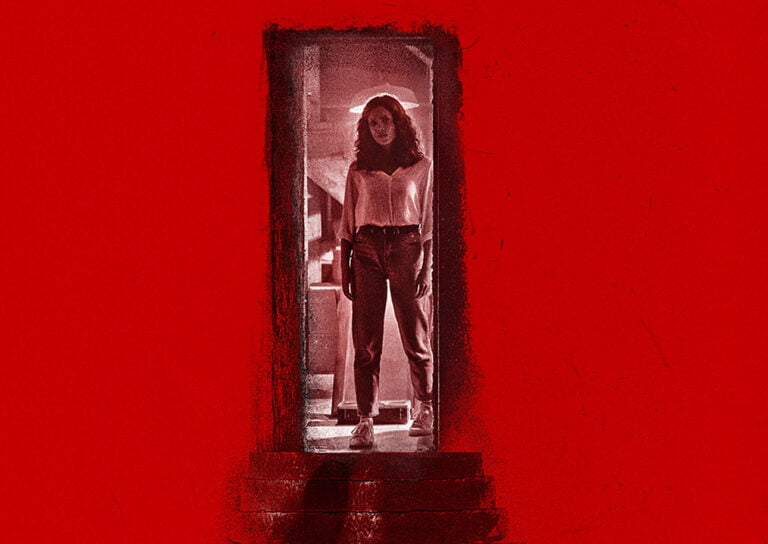 Kobieta w jeansach i białej bluzce stoi w drzwiach na tle czerwonej ściany.