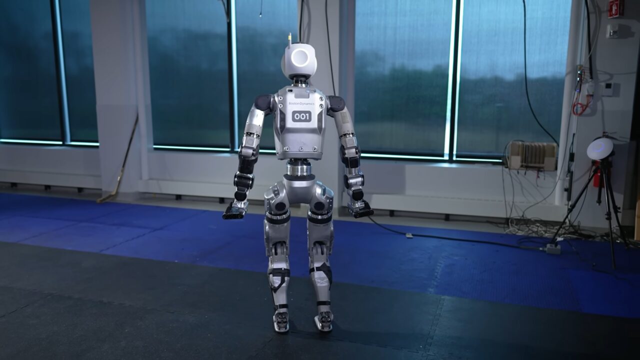 Humanoidalny robot firmy Boston Dynamics stoi na środku pomieszczenia, numer seryjny 001 jest widoczny na jego klatce piersiowej.