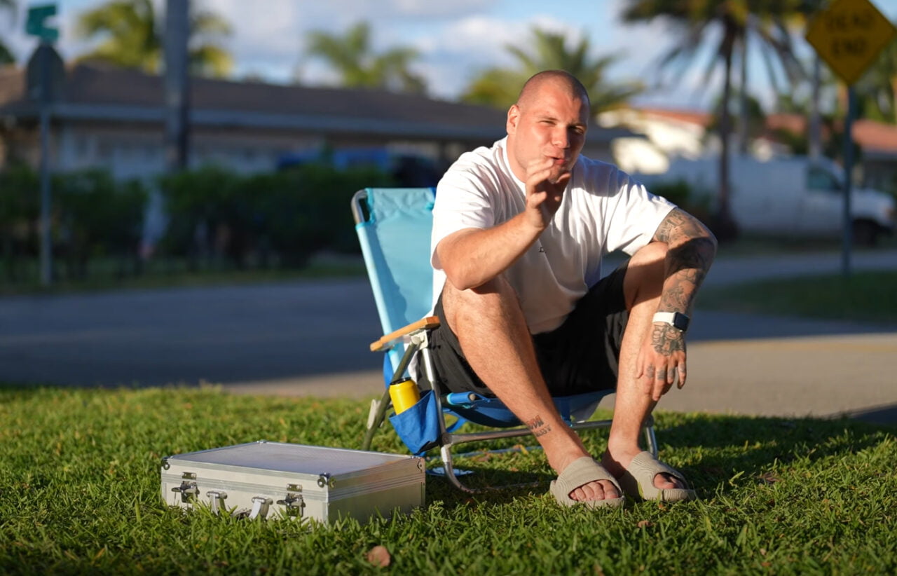 Budda siedzący na składanym krześle na trawniku, machający do kamery, z tatuażami widocznymi na rękach i metalową walizką obok niego.