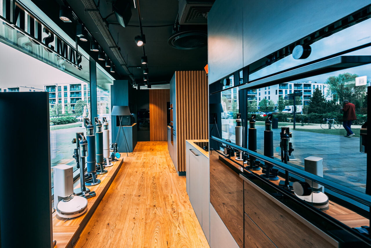 Wnętrze nowoczesnego Cook Story by Samsung  z odkurzaczami, z drewnianą podłogą i dużymi oknami z widokiem na zewnętrzne tereny miejskie.