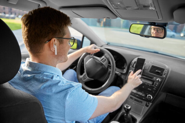 Mężczyzna w okularach i z bezprzewodowymi słuchawkami w uszach prowadzi samochód i patrzy na ekran smartfona.