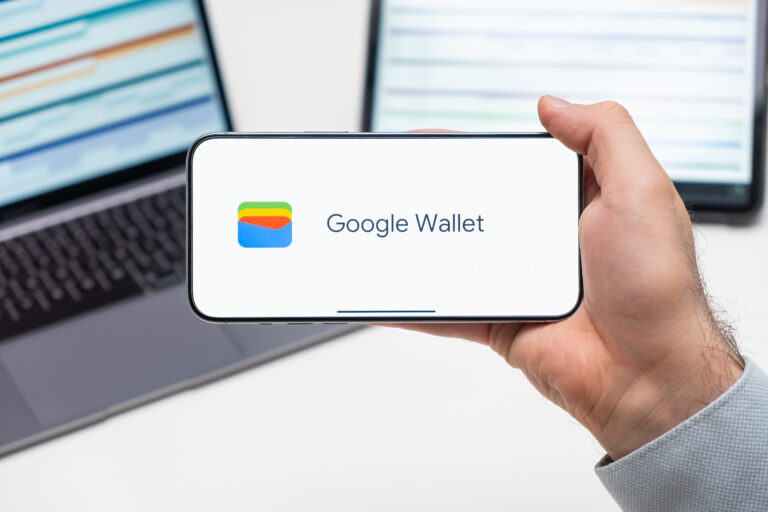 Dłoń trzymająca smartfon z wyświetlonym logotypem Google Wallet, w tle częściowo widoczny laptop.