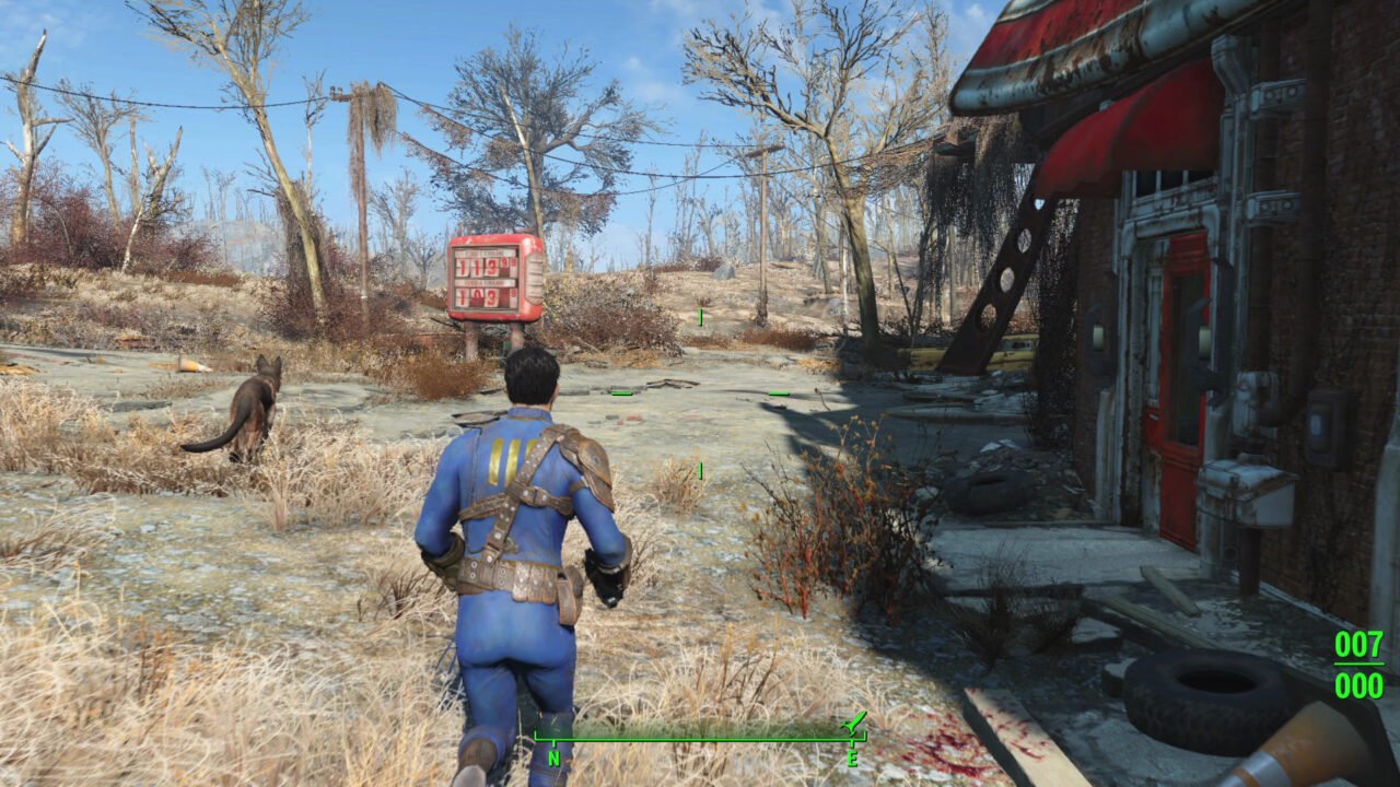 Kadr z gry Fallout 4. Postapokaliptyczny krajobraz z widokiem na plecy postaci gracza w niebieskim kombinezonie i towarzyszącym mu psie, przemierzające opuszczoną ulicę obok zrujnowanej stacji benzynowej, z widocznymi drzewami bez liści i roślinnością w tle.