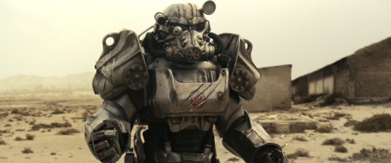Postać z serialu Fallout w pancerzu stojącego na pustynnym terenie z rozmazanym tłem przedstawiającym opuszczone budynki.