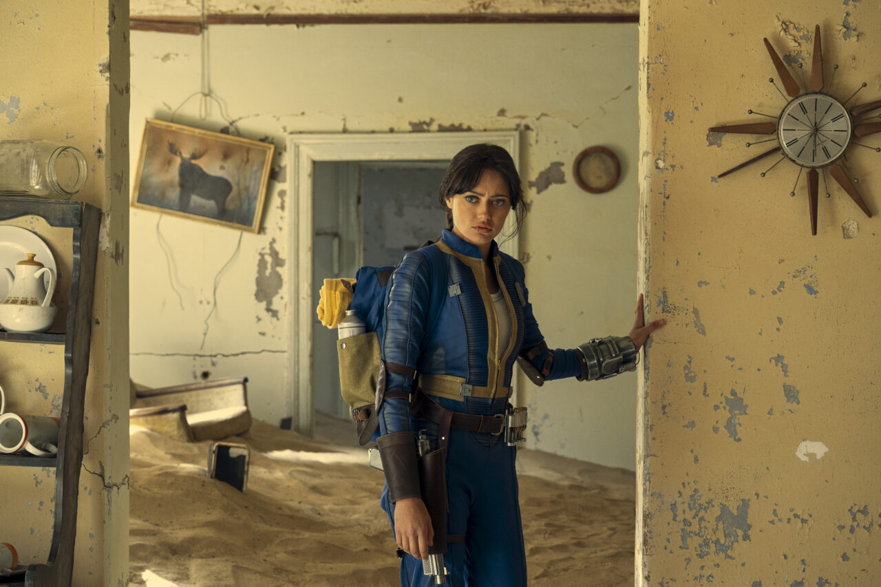 Gdzie obejrzeć Fallout. Kobieta w futurystycznym stroju stoi w opuszczonym pomieszczeniu wypełnionym piaskiem z zawieszoną bronią na przedramieniu.