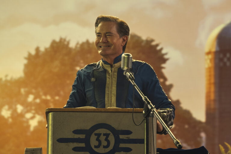 Kadr z serialu Fallout od Amazon Prime. Mężczyzna w średnim wieku ubrany w niebieski strój stojący za mównicą z mikrofonem, z uśmiechem patrzy w bok, w tle zamazane drzewa i wiejski krajobraz o zachodzie słońca.