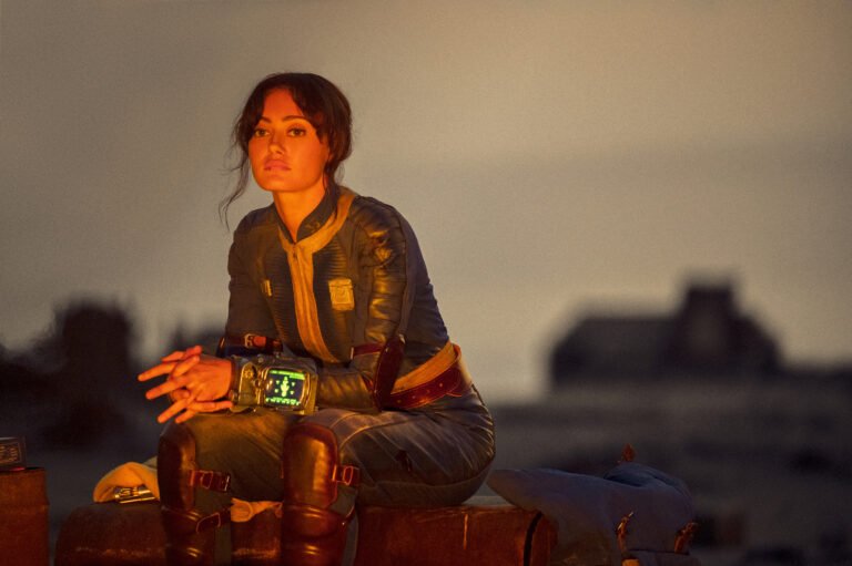 Kobieta w skórzanym kombinezonie pilotki siedzi na walizkach o zmierzchu, z futurystycznym zegarkiem na nadgarstku, w tle nieostra sylwetka budynku. Gdzie obejrzeć Fallout.