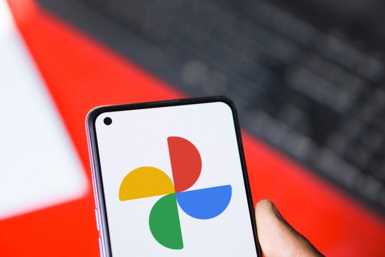 Smartfon trzymany w dłoni z wyświetlonym logo Google Photos na ekranie, na tle klawiatury komputerowej i czerwonej powierzchni.