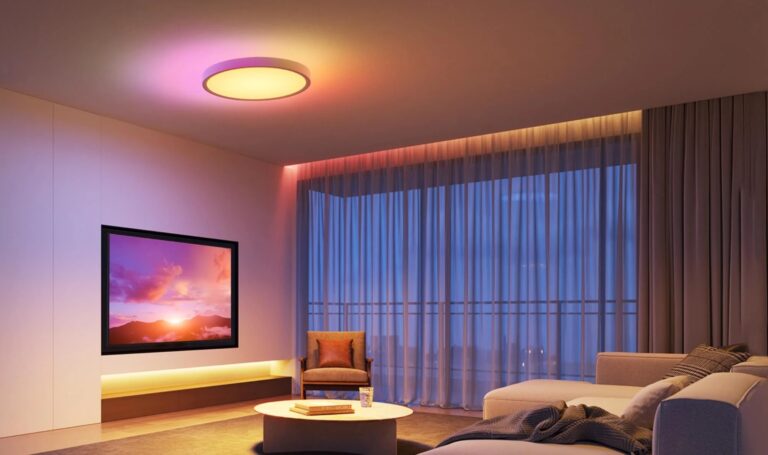 Nowoczesny salon z włączonym telewizorem pokazującym zachód słońca, z miękkim oświetleniem, sofą, fotelikiem i dużymi oknami z zasłonami.