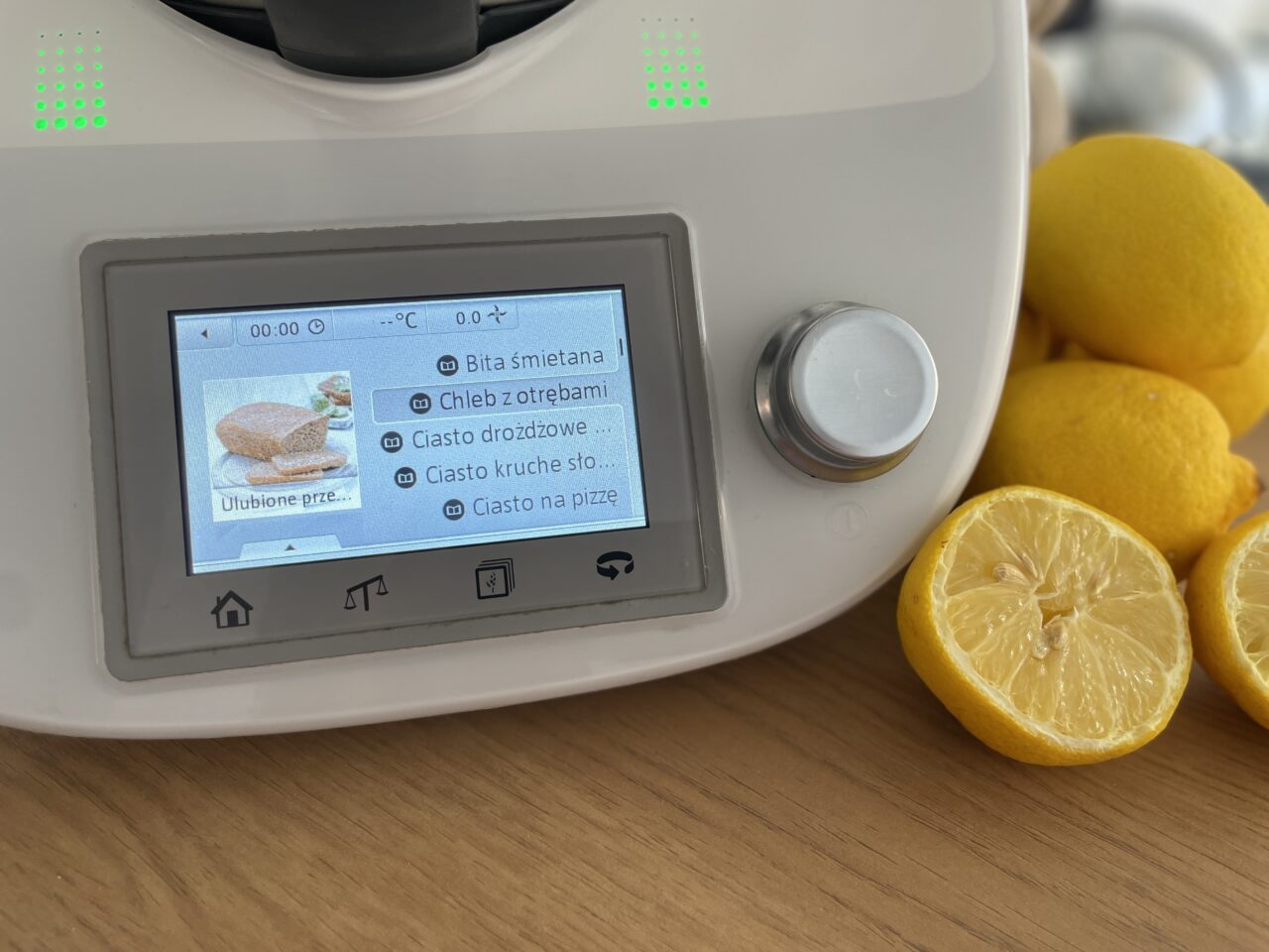 Thermomix TM5. Wyświetlacz wielofunkcyjnego robota kuchennego z wyborem opcji przygotowania jedzenia, w tym bita śmietana i chleb z otrębami, oraz obok leżące cytryny na drewnianym blacie.
