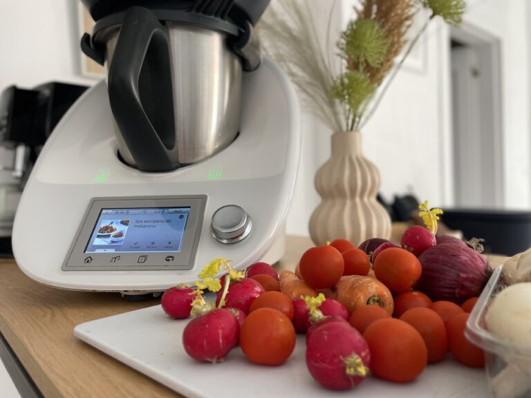 Robot kuchenny na blacie z wyświetlaczem pokazującym przepis na sos, otoczony różnymi warzywami, w tle waza z suchymi trawami.