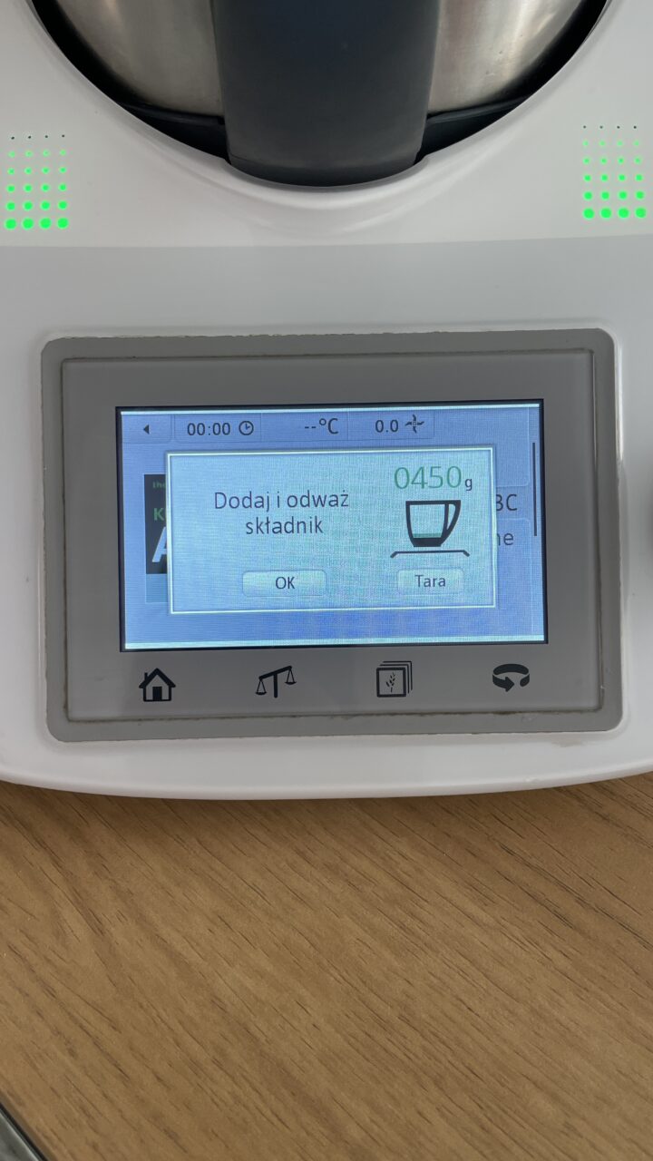 Wyświetlacz urządzenia kuchennego pokazujący komunikat "Dodaj i odważ składnik 0450g" oraz ikonę kubka.
