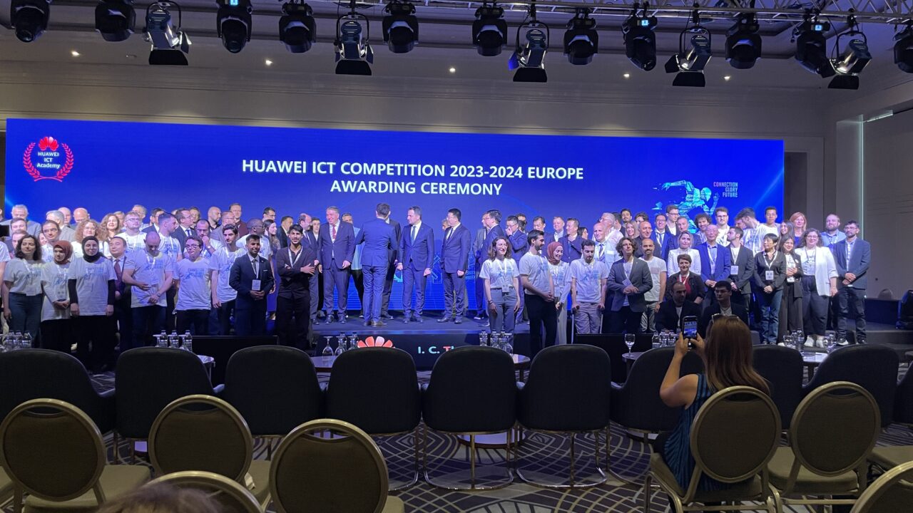Grupa ludzi na scenie podczas ceremonii wręczenia nagród konkursu Huawei ICT 2023-2024 w Europie, sala konferencyjna z krzesłami i oświetleniem scenicznym.