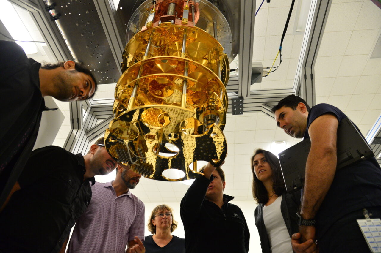 Grupa osób obserwuje duże złote urządzenie naukowe - komputer kwantowy - o złożonej budowie zawieszone w laboratorium.