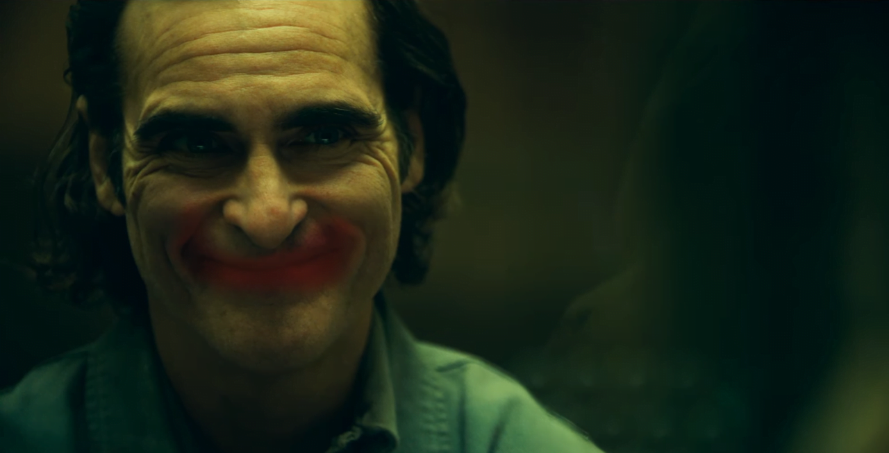 Joker 2. Osoba z namalowaną białą twarzą i czerwonymi ustami, która uśmiecha się na ciemnym tle.