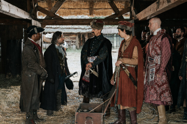 Kadr z filmu Kos. Grupa osób ubranych w historyczne kostiumy rozmawia w drewnianej stodole.