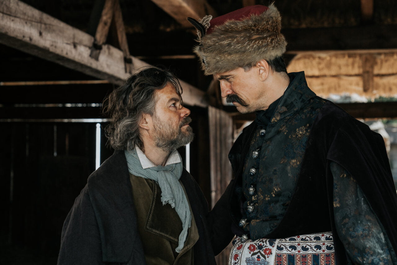 Kadr z filmu Kos. Dwóch mężczyzn w historycznych strojach stoi twarzą w twarz w starej, drewnianej stodole, jeden z nich w futrzanym czapce i zdobnej kurtce, drugi w prostszym ubiorze.
