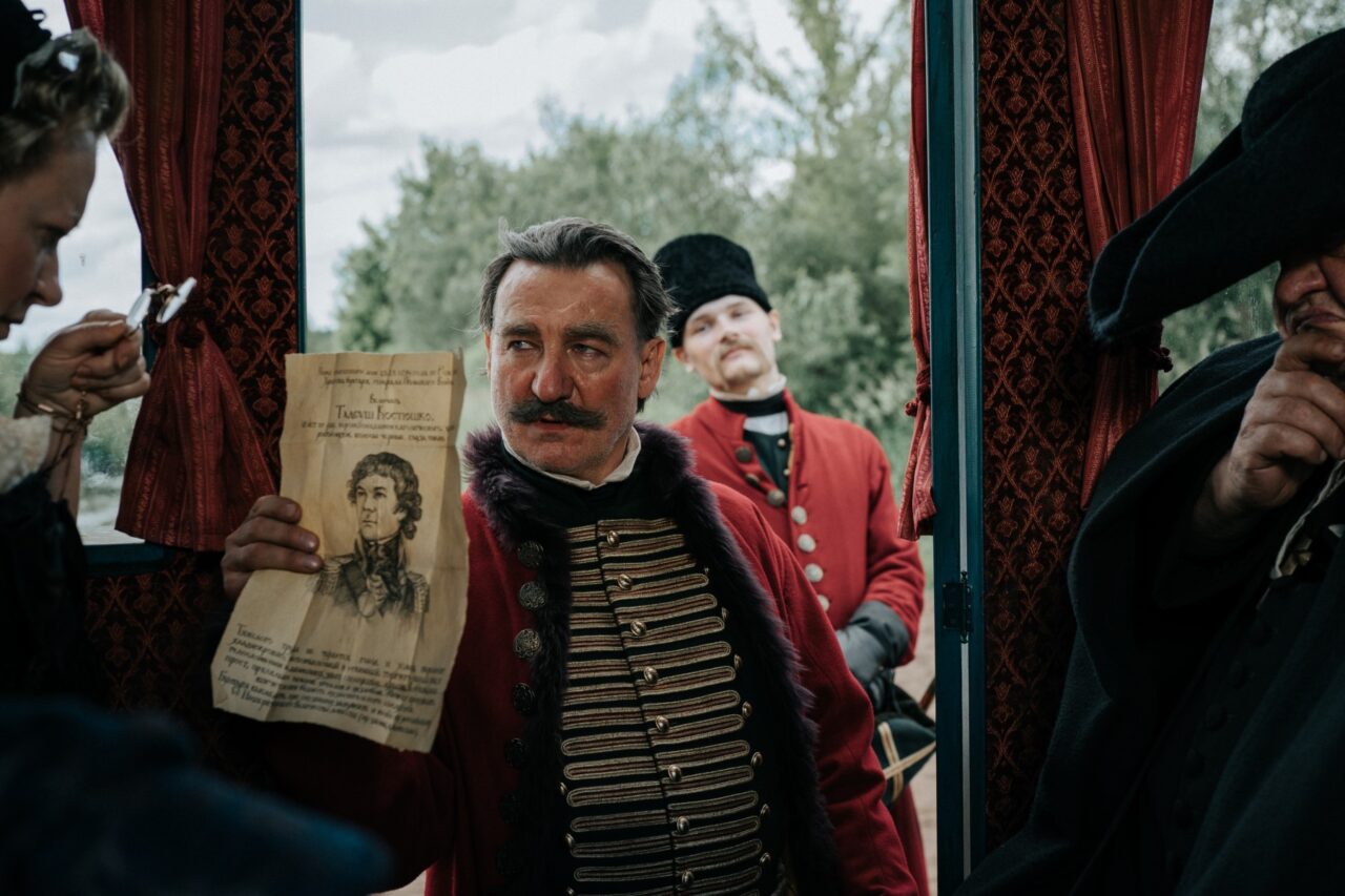 Kadr z filmu Kos. Mężczyzna w historycznym mundurze trzyma przed sobą stary dokument, w tle widoczni inni ludzie w strojach epokowych.