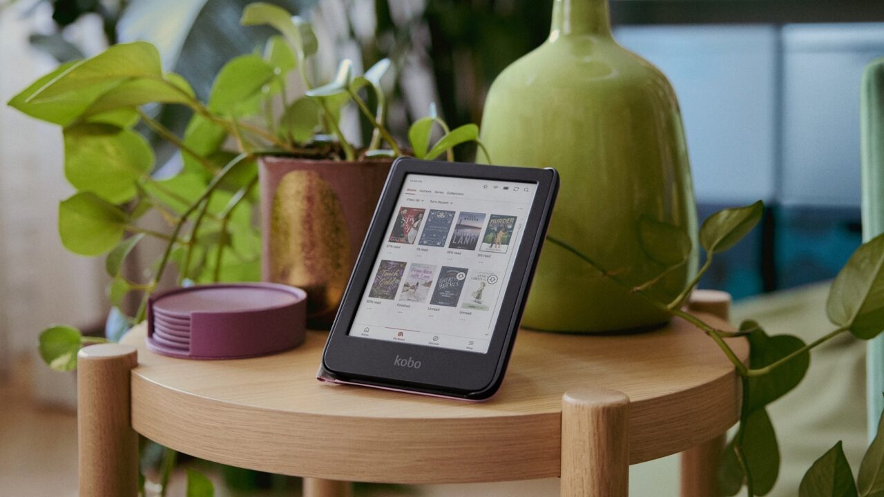 Kolorowy ekran - czytnik e-booków Kobo umieszczony na drewnianym stoliku obok zielonych roślin doniczkowych i dużego zielonego wazonu.