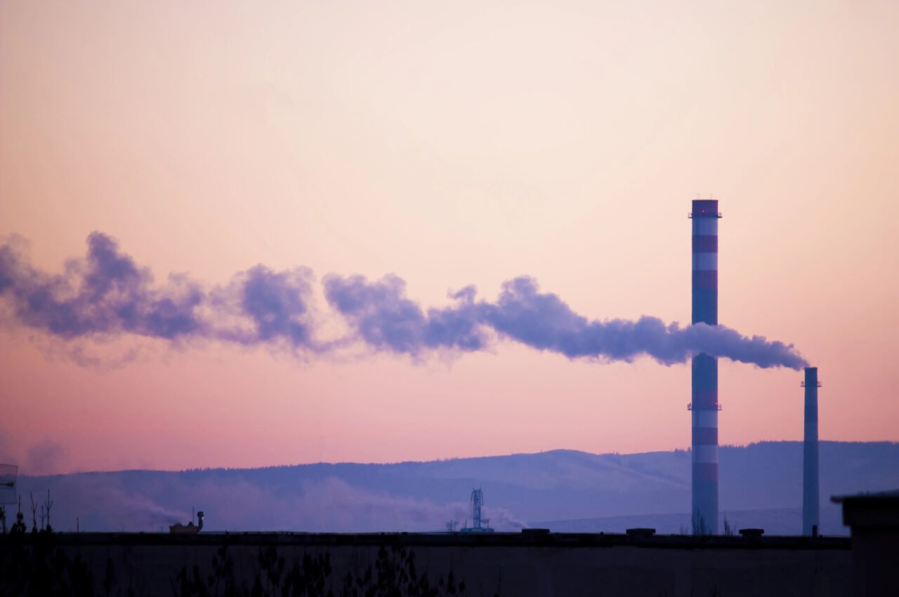 Emisja CO2. Przemysłowe kominy emitujące dym na tle zachodzącego słońca i zarysów gór.